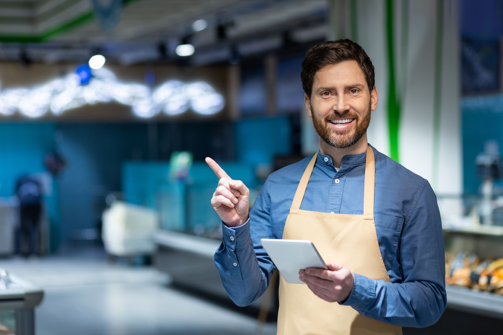 Smiling shop assistant in apron using tablet and pointing at grocery store